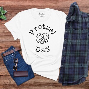 Op de afbeelding: Wit T-shirt met zwarte tekst die "Pretzel Day" zegt en een zwart-witte illustratie van een pretzel.