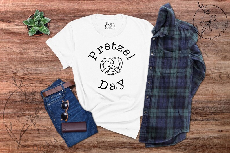 Op de afbeelding: Wit T-shirt met zwarte tekst die "Pretzel Day" zegt en een zwart-witte illustratie van een pretzel.