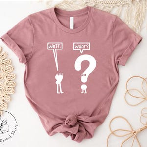 May include: A pink t-shirt with a white graphic of two stick figures. The first stick figure is holding a speech bubble that says "WAIT." The second stick figure is holding a speech bubble that says "WHAT?" and is shaped like a question mark.
