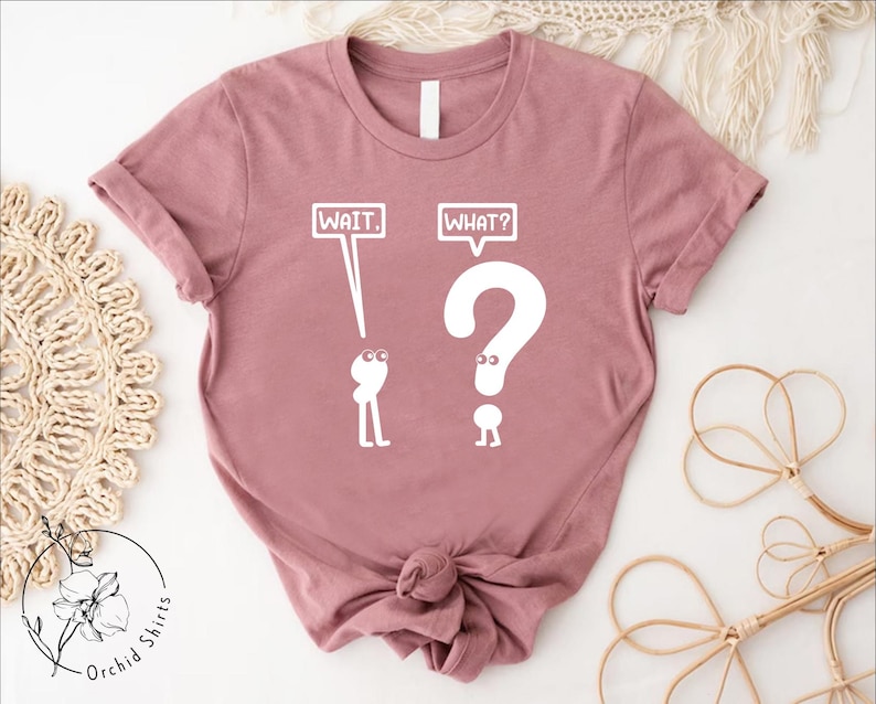 May include: A pink t-shirt with a white graphic of two stick figures. The first stick figure is holding a speech bubble that says "WAIT." The second stick figure is holding a speech bubble that says "WHAT?" and is shaped like a question mark.