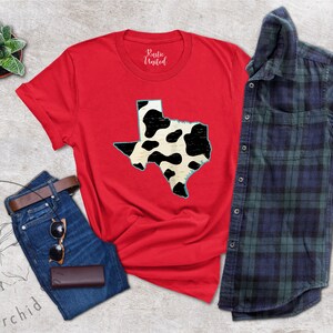 May include: Red t-shirt with a black and white cow print outline of the state of Texas. The shirt is laying flat on a gray surface with a pair of jeans, a small potted plant, and a plaid shirt.