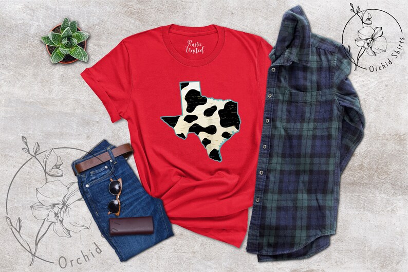 May include: Red t-shirt with a black and white cow print outline of the state of Texas. The shirt is laying flat on a gray surface with a pair of jeans, a small potted plant, and a plaid shirt.