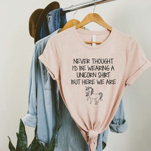 May include: A pink t-shirt with a black graphic that says "Never thought I'd be wearing a unicorn shirt but here we are" with a black and white unicorn illustration.