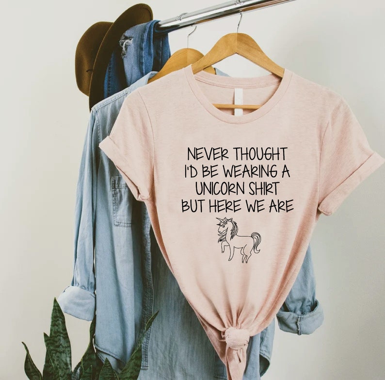May include: A pink t-shirt with a black graphic that says "Never thought I'd be wearing a unicorn shirt but here we are" with a black and white unicorn illustration.