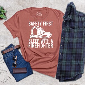 May include: A rust-colored t-shirt with a white graphic of a firefighter helmet and the text "Safety First Sleep with a Firefighter".