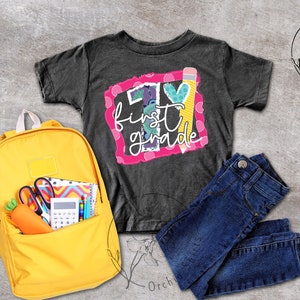 May include: A black t-shirt with a colourful graphic that says "I &hearts; first grade" with a pencil and a heart. A yellow backpack with school supplies is next to the shirt. A pair of blue jeans is also in the image.