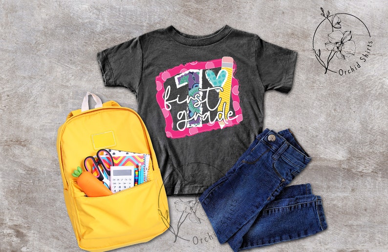 May include: A black t-shirt with a colourful graphic that says "I &hearts; first grade" with a pencil and a heart. A yellow backpack with school supplies is next to the shirt. A pair of blue jeans is also in the image.