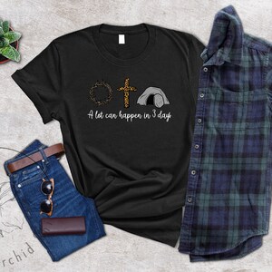 K&ouml;nnte beinhalten: Schwarzes T-Shirt mit einem Aufdruck einer Dornenkrone, einem Kreuz und einem Grab. Der Text "A lot can happen in 3 days" ist unter dem Aufdruck gedruckt.