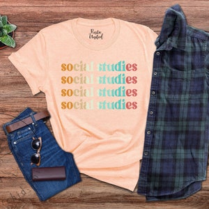 May include: Peach-colored t-shirt with the words "social studies" in a stacked, retro font. The shirt is paired with blue jeans, a brown belt, sunglasses, and a dark brown wallet. A plaid shirt and a small potted succulent are also in the image.