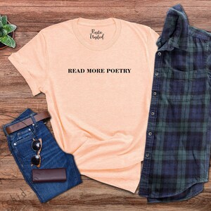 Op de afbeelding: Een perzikkleurig T-shirt met de tekst "READ MORE POETRY" in zwart gedrukt op de voorkant. Het shirt ligt plat op een houten oppervlak met een blauwe spijkerbroek, een bruine leren portemonnee, een zonnebril en een groen-blauw geruit overhemd.