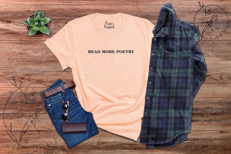 Op de afbeelding: Een perzikkleurig T-shirt met de tekst "READ MORE POETRY" in zwart gedrukt op de voorkant. Het shirt ligt plat op een houten oppervlak met een blauwe spijkerbroek, een bruine leren portemonnee, een zonnebril en een groen-blauw geruit overhemd.