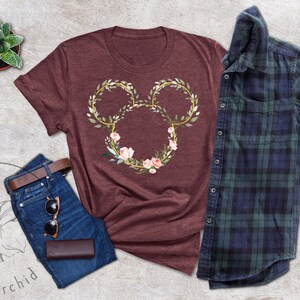 Peut inclure: Un t-shirt bordeaux avec un motif floral de Mickey Mouse. Le motif est compos&eacute; de contours dor&eacute;s, de feuilles vertes et de fleurs roses. Le t-shirt est associ&eacute; &agrave; un jean bleu, une ceinture marron, des lunettes de soleil et une chemise &agrave; carreaux bleue et verte.