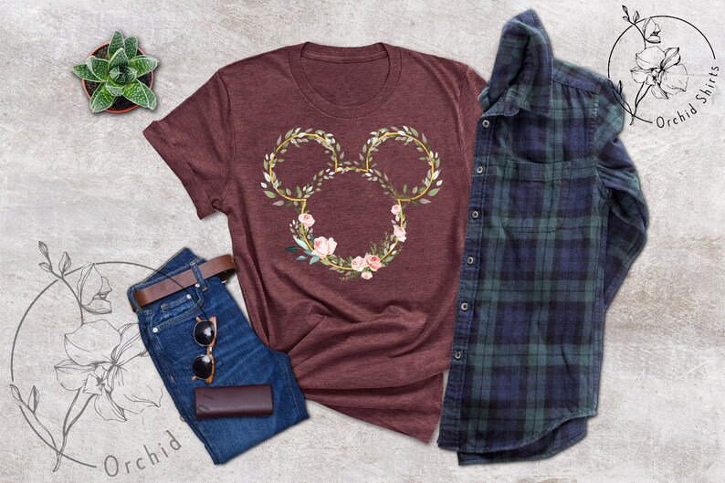 Peut inclure: Un t-shirt bordeaux avec un motif floral de Mickey Mouse. Le motif est compos&eacute; de contours dor&eacute;s, de feuilles vertes et de fleurs roses. Le t-shirt est associ&eacute; &agrave; un jean bleu, une ceinture marron, des lunettes de soleil et une chemise &agrave; carreaux bleue et verte.
