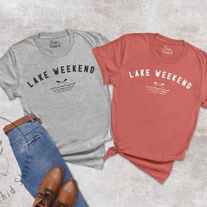 以下が含まれることがあります： グレーとコーラルの2枚のTシャツ。白で「LAKE WEEKEND」の文字がアーチ状に描かれ、交差したオールと波のグラフィックが描かれています。シャツはブルージーンズと茶色の革靴と組み合わせています。
