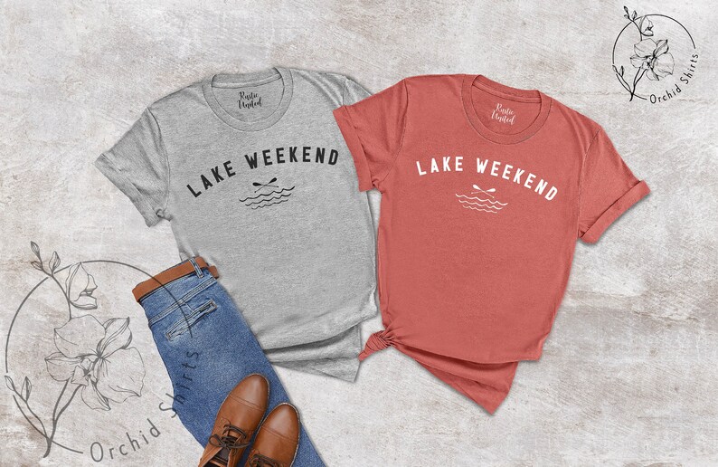 以下が含まれることがあります： グレーとコーラルの2枚のTシャツ。白で「LAKE WEEKEND」の文字がアーチ状に描かれ、交差したオールと波のグラフィックが描かれています。シャツはブルージーンズと茶色の革靴と組み合わせています。