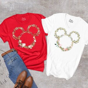 Peut inclure: Deux t-shirts, un rouge et un blanc, chacun avec un motif floral de t&ecirc;te de Mickey Mouse. Le motif est une couronne de feuilles vertes, d'accents dor&eacute;s et de fleurs roses. Les chemises sont associ&eacute;es &agrave; un jean bleu et &agrave; des chaussures marron.