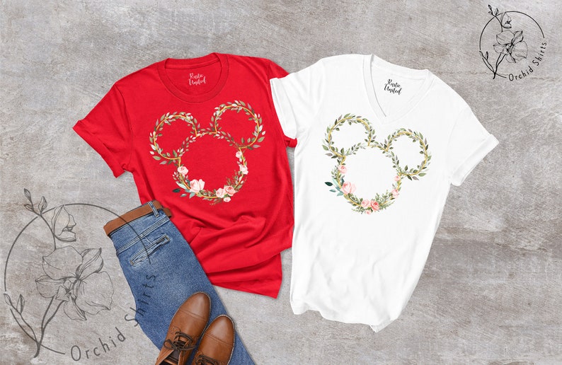 Peut inclure: Deux t-shirts, un rouge et un blanc, chacun avec un motif floral de t&ecirc;te de Mickey Mouse. Le motif est une couronne de feuilles vertes, d'accents dor&eacute;s et de fleurs roses. Les chemises sont associ&eacute;es &agrave; un jean bleu et &agrave; des chaussures marron.