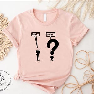 May include: A pink t-shirt with a black graphic of two stick figures. The first stick figure is looking up at the word "WAIT" in a speech bubble. The second stick figure is looking up at a question mark with the word "WHAT?" in a speech bubble.