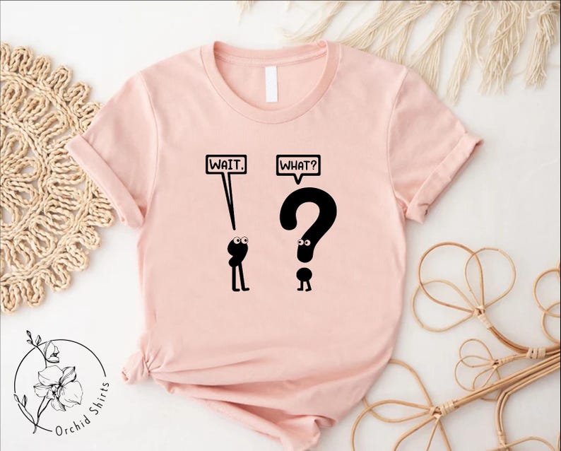 May include: A pink t-shirt with a black graphic of two stick figures. The first stick figure is looking up at the word "WAIT" in a speech bubble. The second stick figure is looking up at a question mark with the word "WHAT?" in a speech bubble.