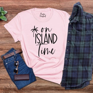 May include: Pink t-shirt with black text that reads "*on island time". The shirt is laid out on a wooden surface with a pair of blue jeans, a brown belt, sunglasses, and a plaid shirt.
