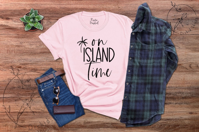 May include: Pink t-shirt with black text that reads "*on island time". The shirt is laid out on a wooden surface with a pair of blue jeans, a brown belt, sunglasses, and a plaid shirt.