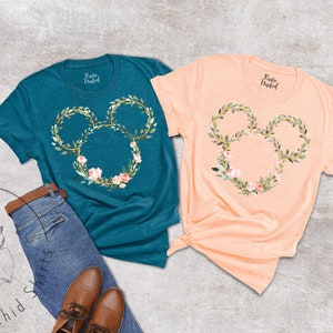 Peut inclure: Deux t-shirts &agrave; manches courtes, un turquoise et un p&ecirc;che, chacun avec un motif floral de t&ecirc;te de Mickey Mouse. Le motif est une couronne de feuilles vertes et de fleurs roses. Les chemises sont pr&eacute;sent&eacute;es avec un jean et des chaussures marron.