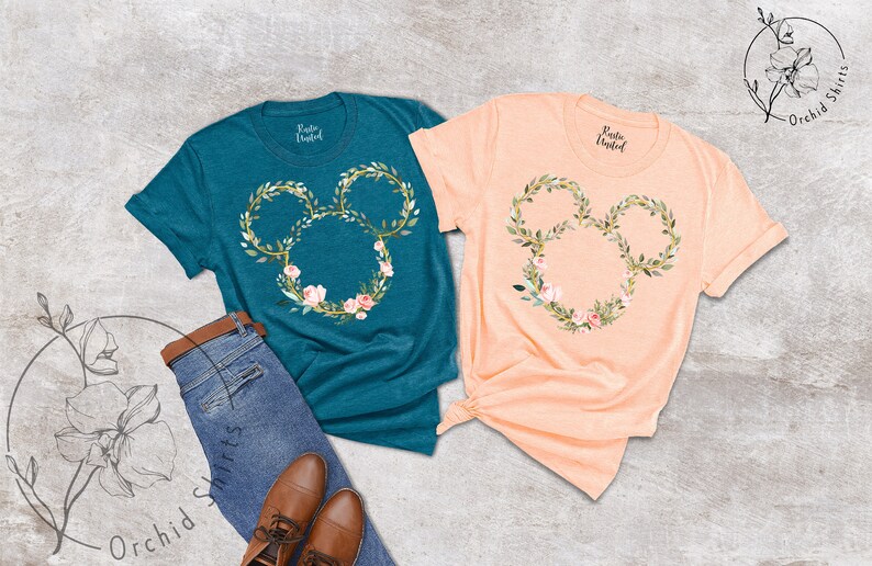 Peut inclure: Deux t-shirts &agrave; manches courtes, un turquoise et un p&ecirc;che, chacun avec un motif floral de t&ecirc;te de Mickey Mouse. Le motif est une couronne de feuilles vertes et de fleurs roses. Les chemises sont pr&eacute;sent&eacute;es avec un jean et des chaussures marron.