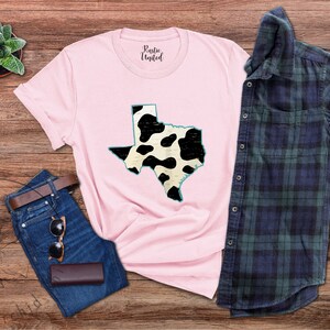 May include: A pink t-shirt with a black and white cow print design of the state of Texas. The shirt is laid out on a wooden surface with a pair of jeans and a plaid shirt.