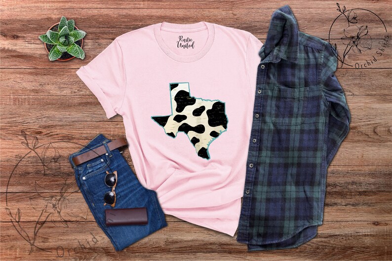 May include: A pink t-shirt with a black and white cow print design of the state of Texas. The shirt is laid out on a wooden surface with a pair of jeans and a plaid shirt.