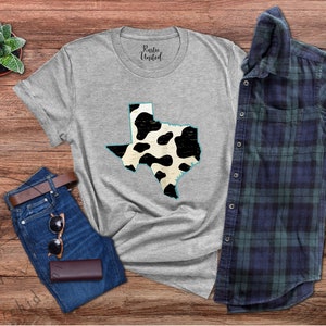 May include: A gray t-shirt with a black and white cow print design of the state of Texas. The shirt is laid flat on a wooden surface with a pair of jeans, sunglasses, and a small potted plant.