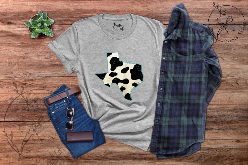 May include: A gray t-shirt with a black and white cow print design of the state of Texas. The shirt is laid flat on a wooden surface with a pair of jeans, sunglasses, and a small potted plant.