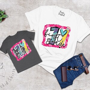 May include: Two t-shirts featuring a graphic design of a large number one with the words "first grade" and a pencil and heart. The shirts are on a grey background with a pair of jeans and sunglasses.