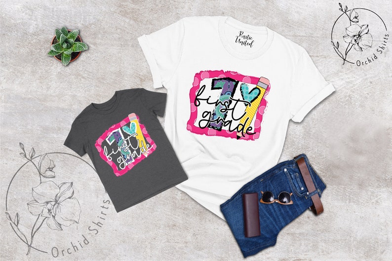 May include: Two t-shirts featuring a graphic design of a large number one with the words "first grade" and a pencil and heart. The shirts are on a grey background with a pair of jeans and sunglasses.