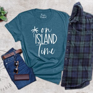 May include: Teal blue t-shirt with white text that reads "on island time" and a palm tree graphic. The shirt is laid flat on a gray textured surface with a pair of blue jeans, a brown leather belt, sunglasses, and a brown leather wallet.