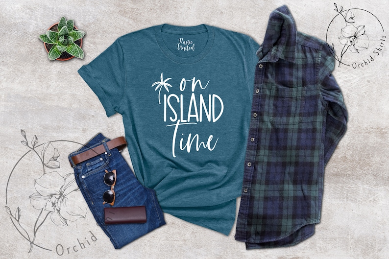 May include: Teal blue t-shirt with white text that reads "on island time" and a palm tree graphic. The shirt is laid flat on a gray textured surface with a pair of blue jeans, a brown leather belt, sunglasses, and a brown leather wallet.