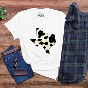 May include: White t-shirt with a black and white cow print design of the state of Texas. The shirt is laid flat on a wooden surface with a pair of jeans, sunglasses, and a plaid shirt.