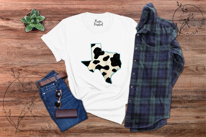 May include: White t-shirt with a black and white cow print design of the state of Texas. The shirt is laid flat on a wooden surface with a pair of jeans, sunglasses, and a plaid shirt.