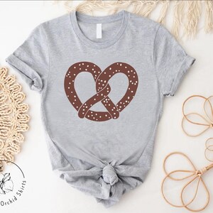 Pu&ograve; includere: Una maglietta grigia con un disegno di pretzel marrone. Il pretzel ha delle spruzzate bianche.