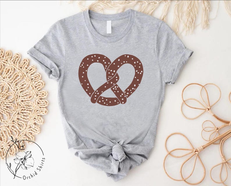 Pu&ograve; includere: Una maglietta grigia con un disegno di pretzel marrone. Il pretzel ha delle spruzzate bianche.