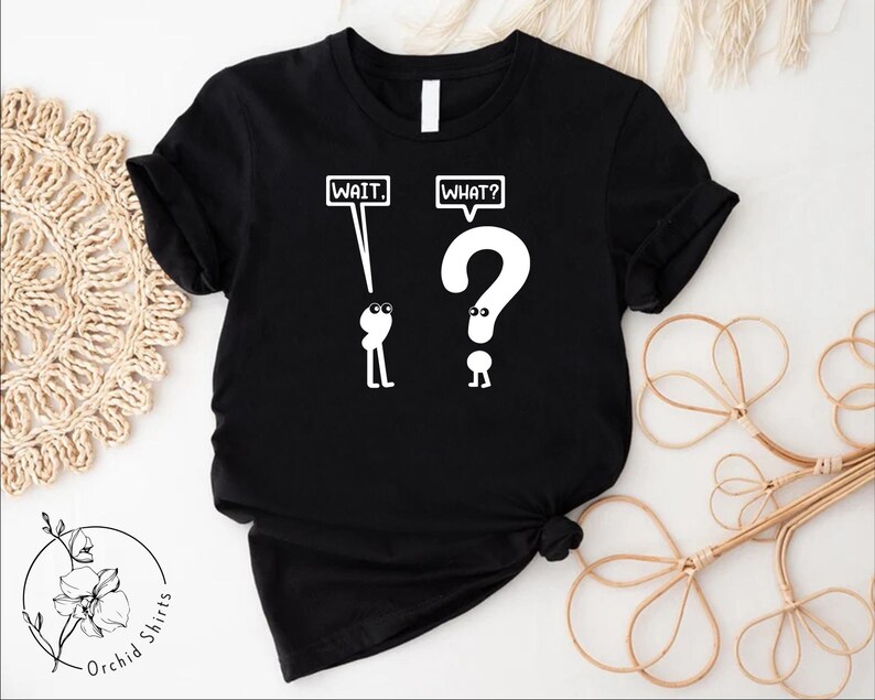 May include: Black t-shirt with a white graphic of two stick figures. The figure on the left holds a sign that says "WAIT." The figure on the right holds a question mark and a sign that says "WHAT?"