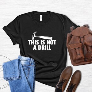 May include: Black t-shirt with a white hammer and the text "This is not a drill". The shirt is laid out on a wooden surface with a pair of blue jeans, a brown leather backpack, and brown leather boots.