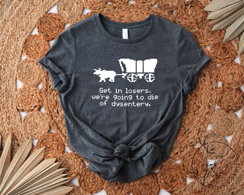 Pu&ograve; includere: T-shirt grigio scuro con un design pixelato di un carro e un bue, con la scritta "Get in losers, we're going to die of dysentery." La maglietta &egrave; annodata in basso, appoggiata su un tappeto rotondo tessuto.