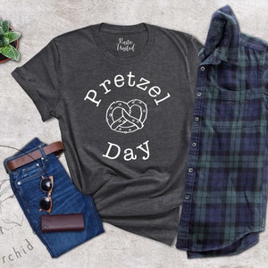 Op de afbeelding: Een donkergrijs T-shirt met de tekst "Pretzel Day" en een witte omtrek van een pretzel. Het shirt ligt op een lichtgrijze ondergrond met een spijkerbroek, een leren portemonnee, een zonnebril en een potplant.