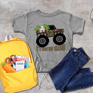 May include: A grey t-shirt featuring a monster truck graphic with a bunny and carrots, and the text "It's Hunting Season!". The image also shows a yellow backpack, blue jeans, and school supplies.