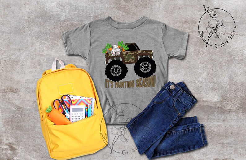 May include: A grey t-shirt featuring a monster truck graphic with a bunny and carrots, and the text "It's Hunting Season!". The image also shows a yellow backpack, blue jeans, and school supplies.