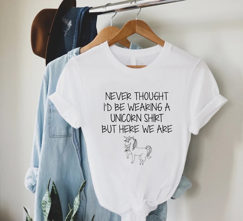 May include: White t-shirt with a black graphic of a unicorn and the text "Never thought I'd be wearing a unicorn shirt but here we are."