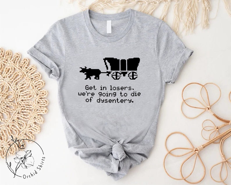 Pu&ograve; includere: T-shirt grigio chiaro con un'immagine pixelata nera di un carro coperto trainato da un orso. Il testo "Get in losers, we're going to die of dysentery" &egrave; stampato sotto. La maglietta &egrave; di Orchid Shirts.