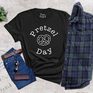 Op de afbeelding: Zwart T-shirt met witte tekst die "Pretzel Day" zegt en een witte omtrek van een pretzel.