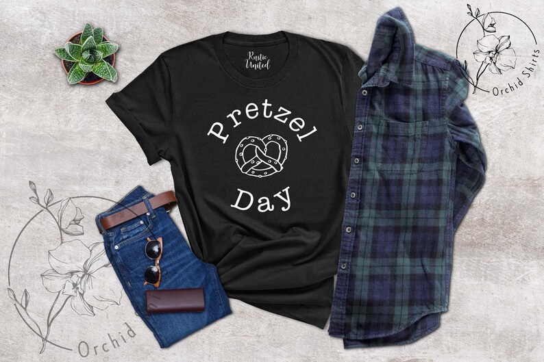 Op de afbeelding: Zwart T-shirt met witte tekst die "Pretzel Day" zegt en een witte omtrek van een pretzel.