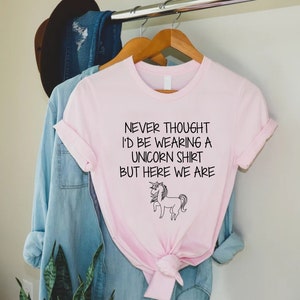 May include: Pink t-shirt with a black unicorn graphic and the text "Never thought I'd be wearing a unicorn shirt but here we are."